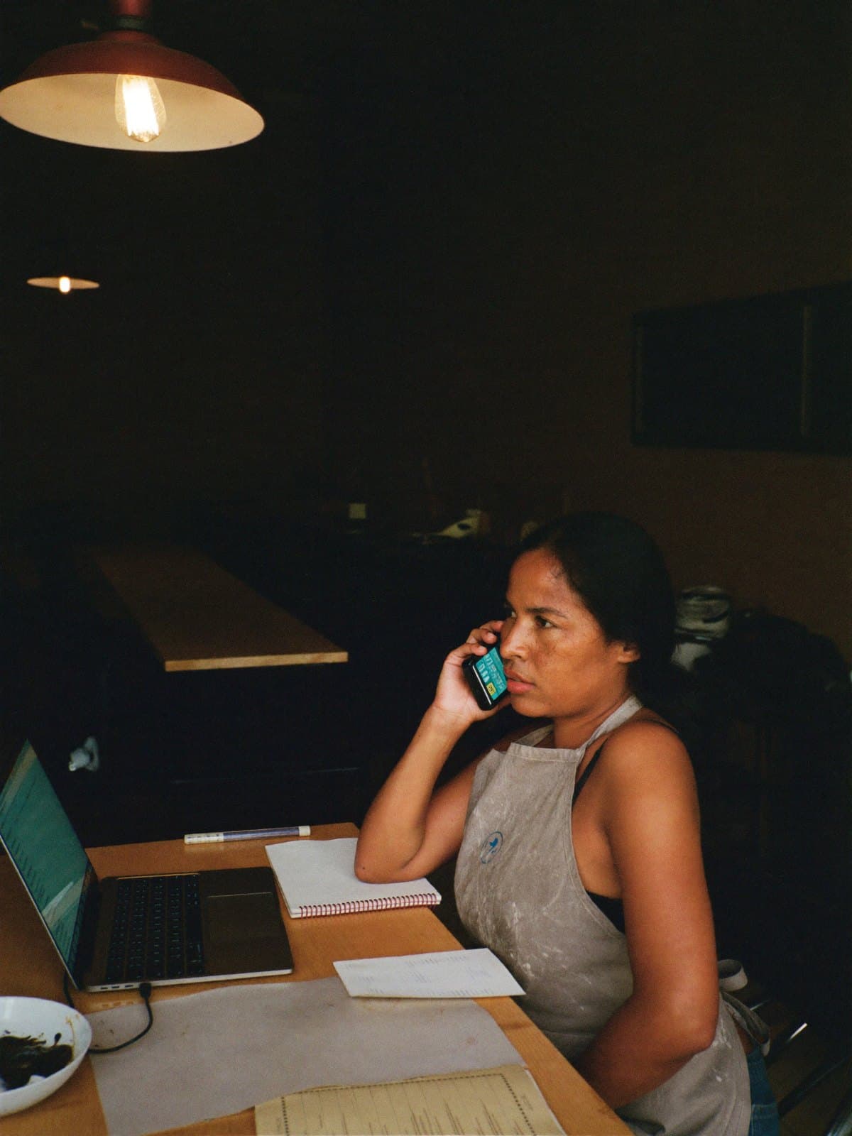 A woman in an apron on the phone at a desk after close, a laptop and a notebook beside her