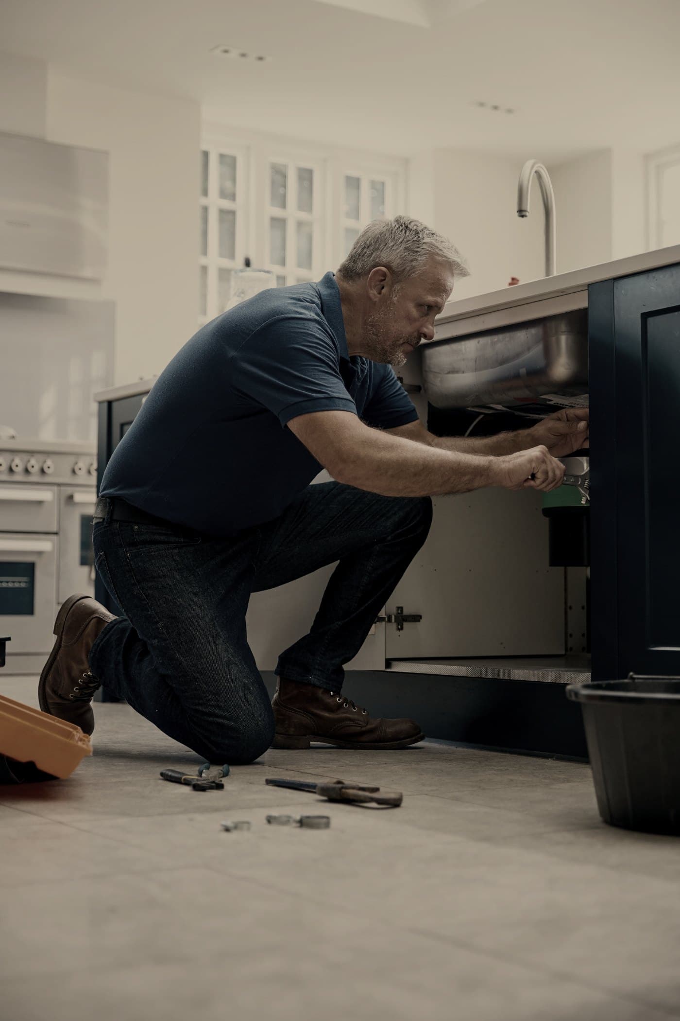 A plumber kneeling at an open kitchen cabinet, reaching in under the sink